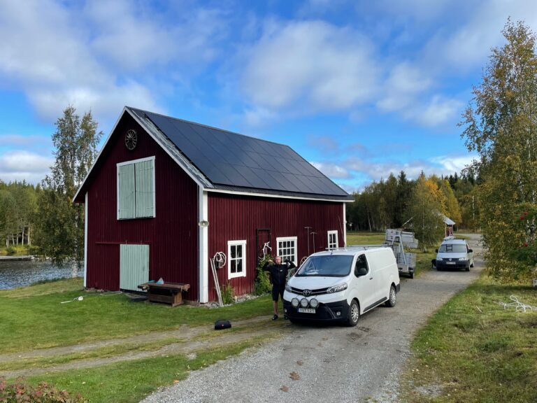 Ett rött hus med solpaneler på taket vid en väg med parkerade bilar på en solig dag.